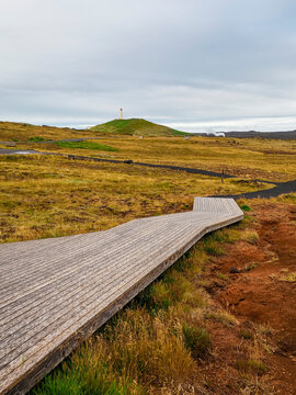Wild Icelandic landscape with lava fields, local vegetation, wooden track, black pebble trail near Gunnuhver Hot Springs. In the distance visible Reykjanesviti Lighthouse. Reykjanes Peninsula, Iceland