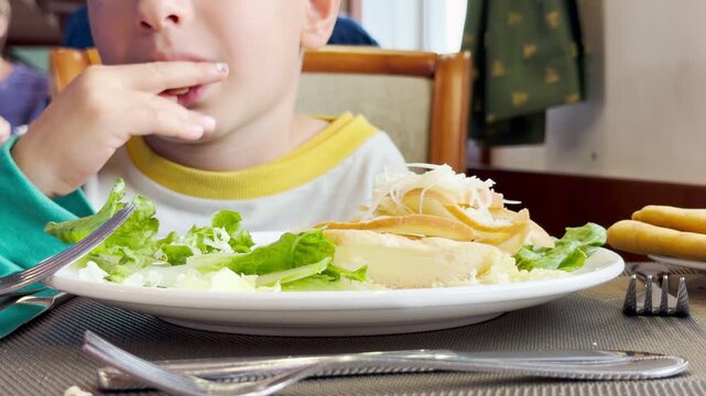 A happy boy eats a tasty meal at a dining table. He enjoys his fresh salad with content curiosity inside a cozy cruise ship restaurant.