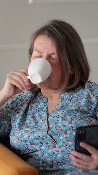 Woman uses smartphone beside white mug in relaxed home setting. She glances at screen, then drinks from cup, keeping both phone and beverage close during rest while seated in chair and holding mug.