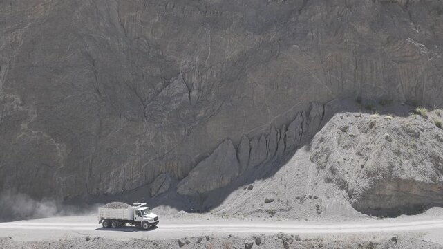 Aerial view of a loaded dump truck driving along a dusty dirt road in a sand and gravel quarry beneath a towering rock wall, D-Log M