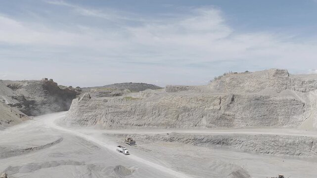 Aerial panoramic view of an open pit sand and gravel quarry with dump trucks and heavy machinery on dusty haul roads under a blue sky while the camera flies backward, D-Log M