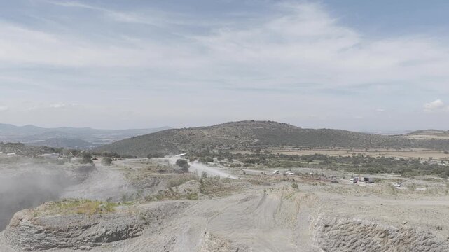 Aerial reveal shot of a quarry rock wall as the camera ascends vertically to unveil a wide valley with farmland, hills and a distant city under a blue sky, D-Log M