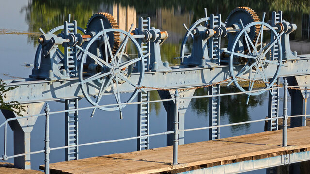 Blue metal wheels and gears of an old water control dam mechanism above a calm river with reflections and a wooden walkway
