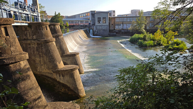 Industrial river dam with concrete pillars and blue sluice gates surrounded by green trees and old factory buildings