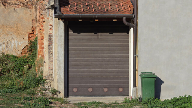 Closed dark brown sectional garage door between weathered brick wall and grey concrete wall with green wheelie bin