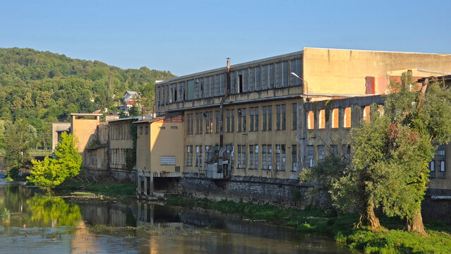 Old weathered industrial factory building standing on the banks of a calm river surrounded by lush green trees and forest hills under a clear blue sky