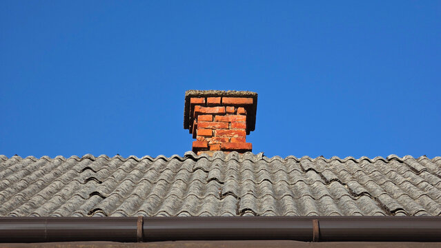 Old red brick chimney on a corrugated gray roof under a clear blue sky with a brown gutte