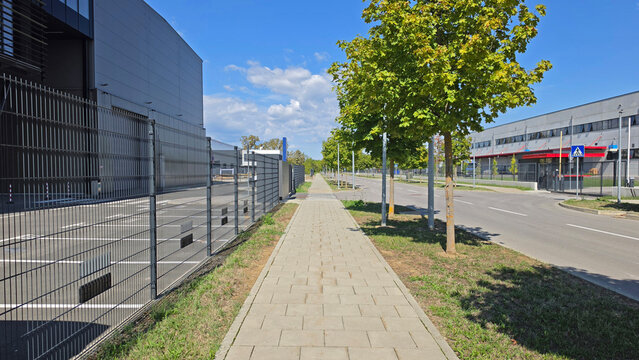 Empty paved pedestrian walkway beside an asphalt street and metal fencing in a modern industrial commercial park on a sunny day
