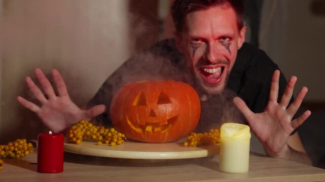 Man in joker look mystically appears near jack o lantern, grins, gives frightening stare at camera and walks away. Makeup, character for halloween night, carnival transformations into scary figures.