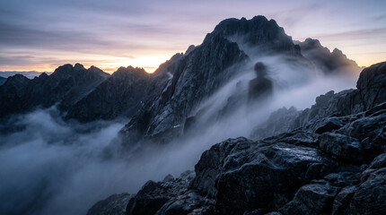Obraz premium Silhouette of a person standing on a rocky mountain peak above clouds at sunset.