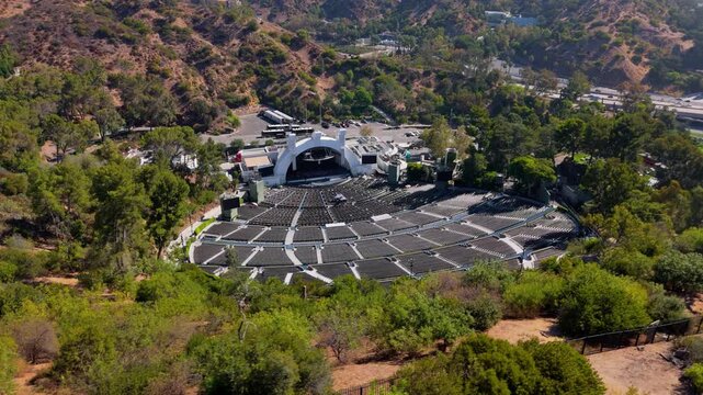 Aerial view of famous open air amphitheater in Hollywood Hills. Empty seating rows and white stage shell surrounded by green trees. Sunny summer day in Los Angeles, California. 