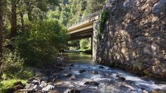 A concrete road bridge with iron railing spans a clear rocky mountain stream, flanked by a stone retaining wall covered in ivy, surrounded by lush green forest trees
