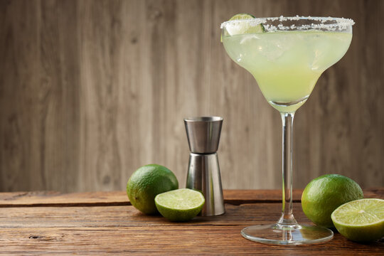 Refreshing margarita cocktail in glass, limes and jigger on wooden table, closeup. Space for text
