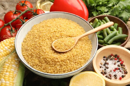 Raw bulgur in bowl, spoon and other products on wooden table, closeup