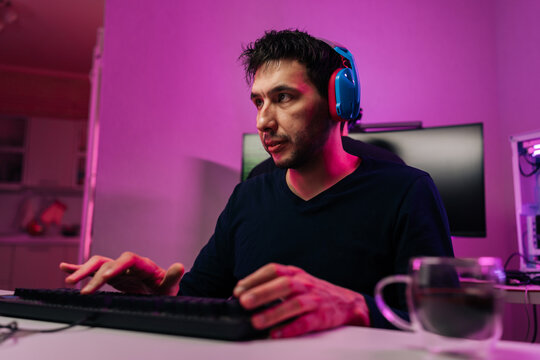 Focused man with headset plays computer game at night. face showing intense concentration. Modern gaming setup with vibrant pink neon lighting creates futuristic atmosphere. Hobby esports concept.