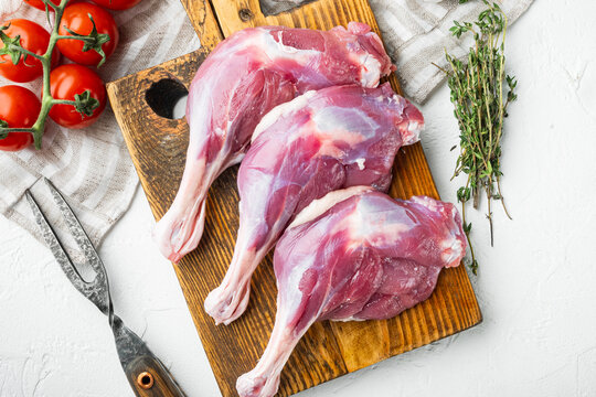 Three raw duck leg thighs with visible bone and fat marbling arranged on a rustic wooden cutting board beside fresh thyme sprigs and vine tomatoes, metal fork with textured handle to the side