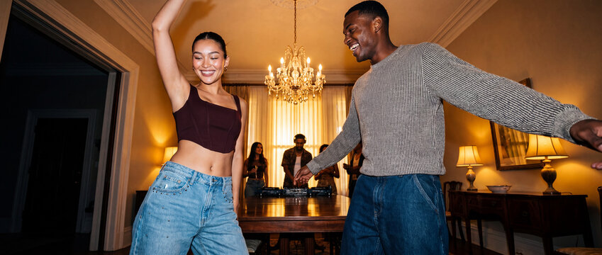 Friends dancing at home party with smiling young multicultural couple moving to music under a chandelier while DJ plays in the background, perfect for social lifestyle and celebration ads
