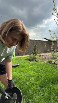 A woman with long blonde hair is engaged in yard work, holding a garden tool near freshly cut green grass under a cloudy sky, suggesting outdoor activity in a backyard setting