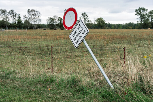 Leaning traffic sign with German text &ldquo;Land und forstwirtsch Verkehr frei (Agricultural and forestry traffic allowed)&rdquo; beside wire fence in rural grassy landscape under cloudy sky.