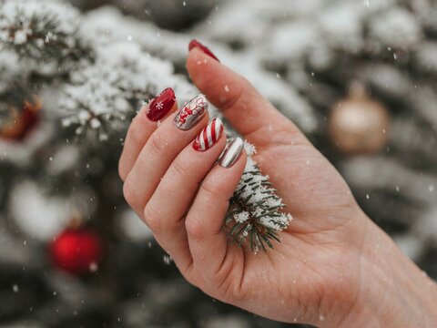 Festive manicure with jewel-toned red and silver nails, captured close-up against blurred snow-covered branches and Christmas decorations.