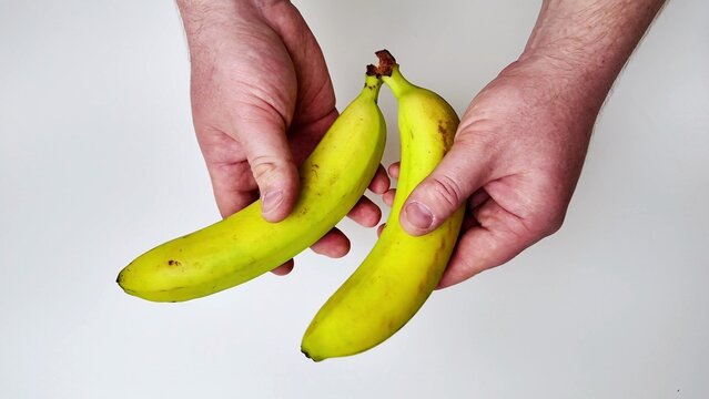 Two hands hold two yellow bananas against a plain white background, with the fingers gripping the fruit from opposite sides
