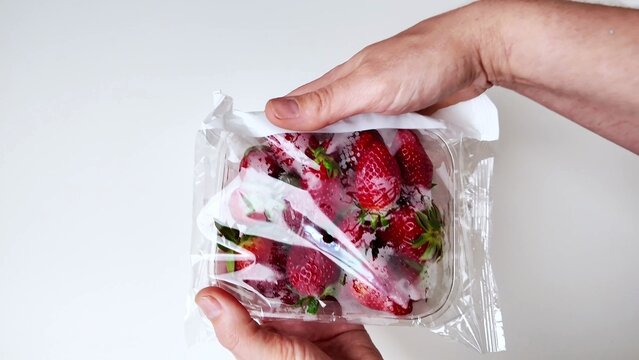 hands hold a clear plastic container of red strawberries wrapped in a transparent plastic bag, placed against a white background