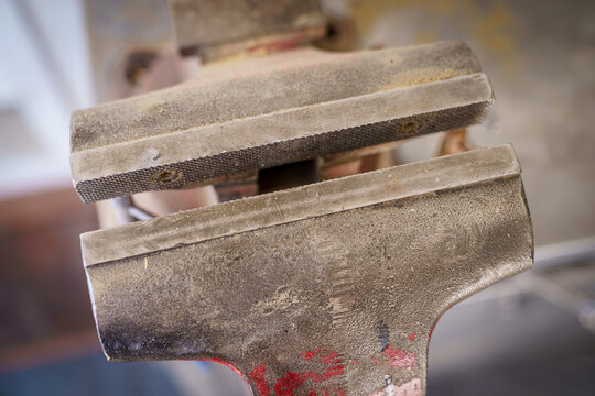 Detailed close-up of an aged metal bench vise with textured jaws, worn surfaces, dust, scratches, and traces of red paint, highlighting the rugged appearance of a well-used workshop tool.
