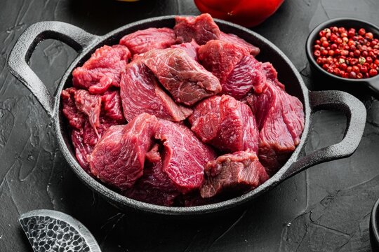 Diced raw beef chunks arranged in a black cast iron frying pan on a dark textured stone surface beside red peppercorns and a slice of red bell pepper under directional lighting