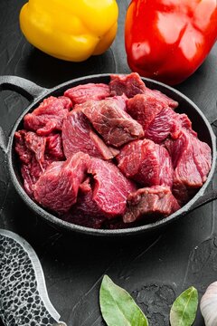 Chopped raw lean beef chunks in a black cast iron skillet beside whole red and yellow bell peppers on dark stone surface with bay leaves and garlic clove under directional light