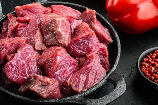 Chopped raw beef chunks in a cast iron frying pan beside a whole red bell pepper and a small bowl of pink peppercorns on a dark textured stone surface under soft directional lighting