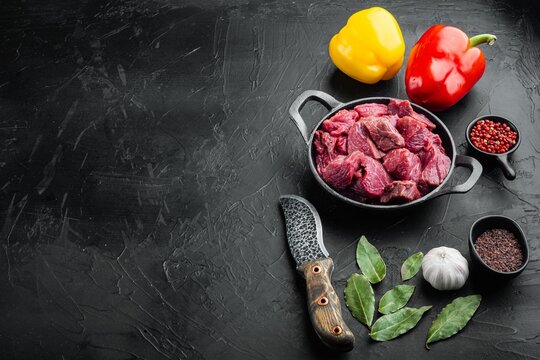 Red and yellow bell peppers capsicum beside diced raw beef chunks in a cast iron pan with garlic cloves bay leaves red and black peppercorns and a patterned knife on a dark textured stone surface