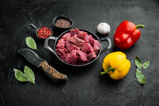 Red and yellow sweet bell peppers arranged beside a cast iron frying pan filled with raw chopped beef chunks, whole garlic bulb, bay leaves, small bowls of black and red peppercorns