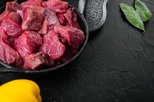 Bay leaves resting beside raw beef chuck chunks in a cast iron frying pan on textured black stone surface with yellow sweet pepper and copy space under directional light