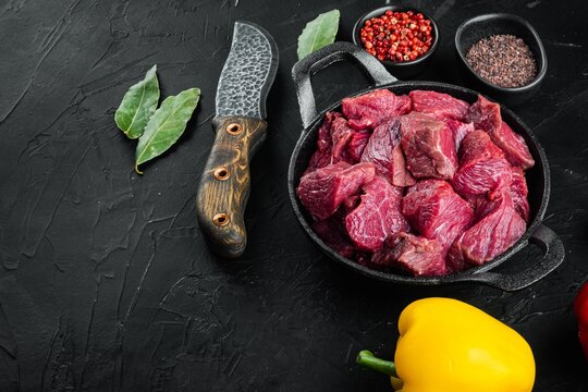Yellow and red sweet bell peppers beside chopped raw beef chunks in a cast iron frying pan with bay leaves peppercorns and red chili on black textured stone surface under directional light