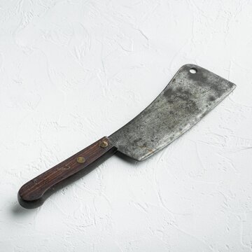 Weathered metal blade of a butcher cleaver with dark wooden handle secured by brass rivets resting on textured white stone surface under even diffused light