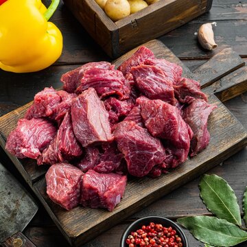 Diced raw beef chunks arranged on a dark rustic wooden cutting board beside whole yellow bell pepper and fresh bay leaves, small black bowl of red peppercorns nearby