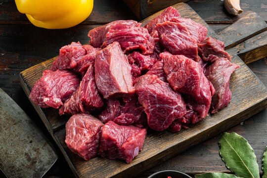 Diced raw stewing beef cubes arranged on a dark weathered wooden cutting board beside whole yellow bell pepper and fresh bay leaves under warm directional light