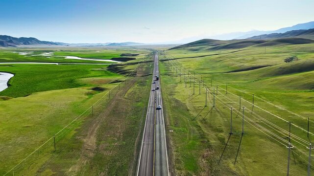 High angle view of a straight asphalt highway stretching across green grasslands and rolling hills under a blue sky in Xinjiang, China.