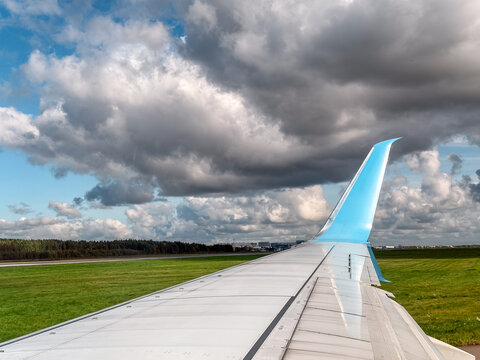 Passenger plane airplane wing view from a window with a blue upward winglet tip on an airport runway ground. Beautiful clear summer sky background template with dramatic white cumulus clouds. 