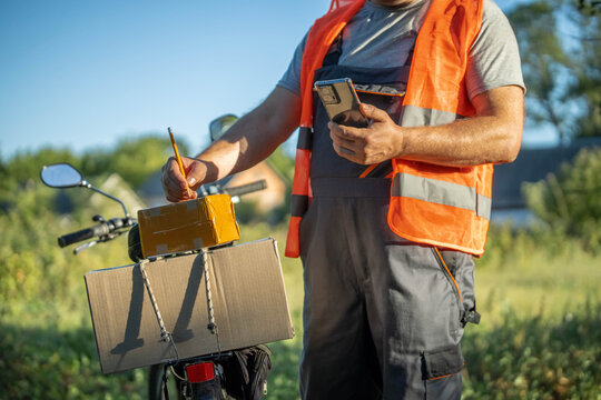 A delivery man in an orange vest stands by his bicycle, checking his device as he organizes packages in a park. The sun shines brightly, creating a cheerful atmosphere