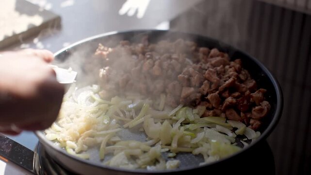 Person adds chopped garlic to frying pan with cooked meat and sliced onion. Concept of seasoning food and home cooking