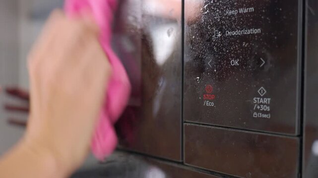 Closeup of wet microwave control panel as hand wipes away cleaner with pink cloth. Concept of appliance maintenance, surface cleaning and kitchen hygiene