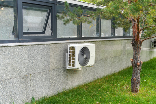 White air conditioner outdoor unit mounted on grey stone exterior wall under window. HVAC climate control equipment. Real Photo