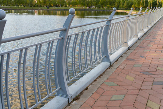 Curved grey metal handrail along tiled walkway next to water lake in public park. Exterior. Real Photo