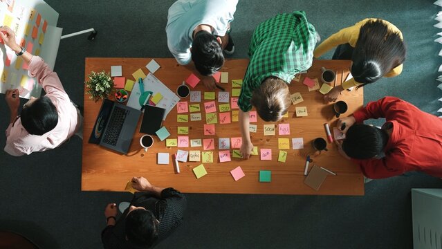 Top view of skilled business people discuss plan at meeting room while writing idea at sticky notes and stick at whiteboard. Group of diverse team working together to planning strategy. Convocation.