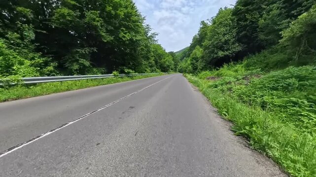 First-person POV driving shot on an asphalt mountain road passing lush green forest hills with a oncoming white car and dramatic cloud formations overhead.