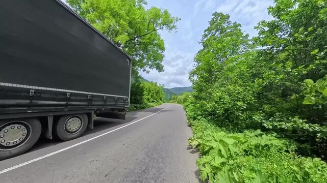 First-person POV driving shot on an asphalt mountain road passing lush green forest hills with a oncoming white car and dramatic cloud formations overhead.