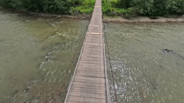First-person POV perspective walking across a narrow wooden suspension footbridge spanning over a wide, fast-flowing mountain river toward the green forest shore.
