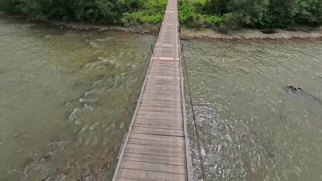 A wooden suspension bridge spanning a river or watercourse. A bridge consists of a deck made of planks. First-person POV perspective walking across a narrow wooden suspension footbridge 