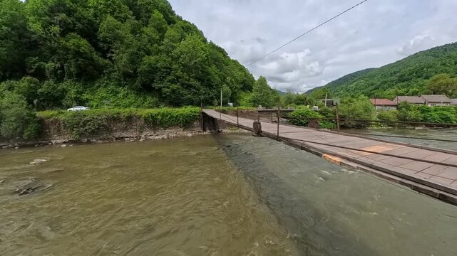 First-person POV perspective looking across a narrow wooden suspension bridge crossing a wide, flowing mountain river towards lush green forested hills under a cloudy sky.